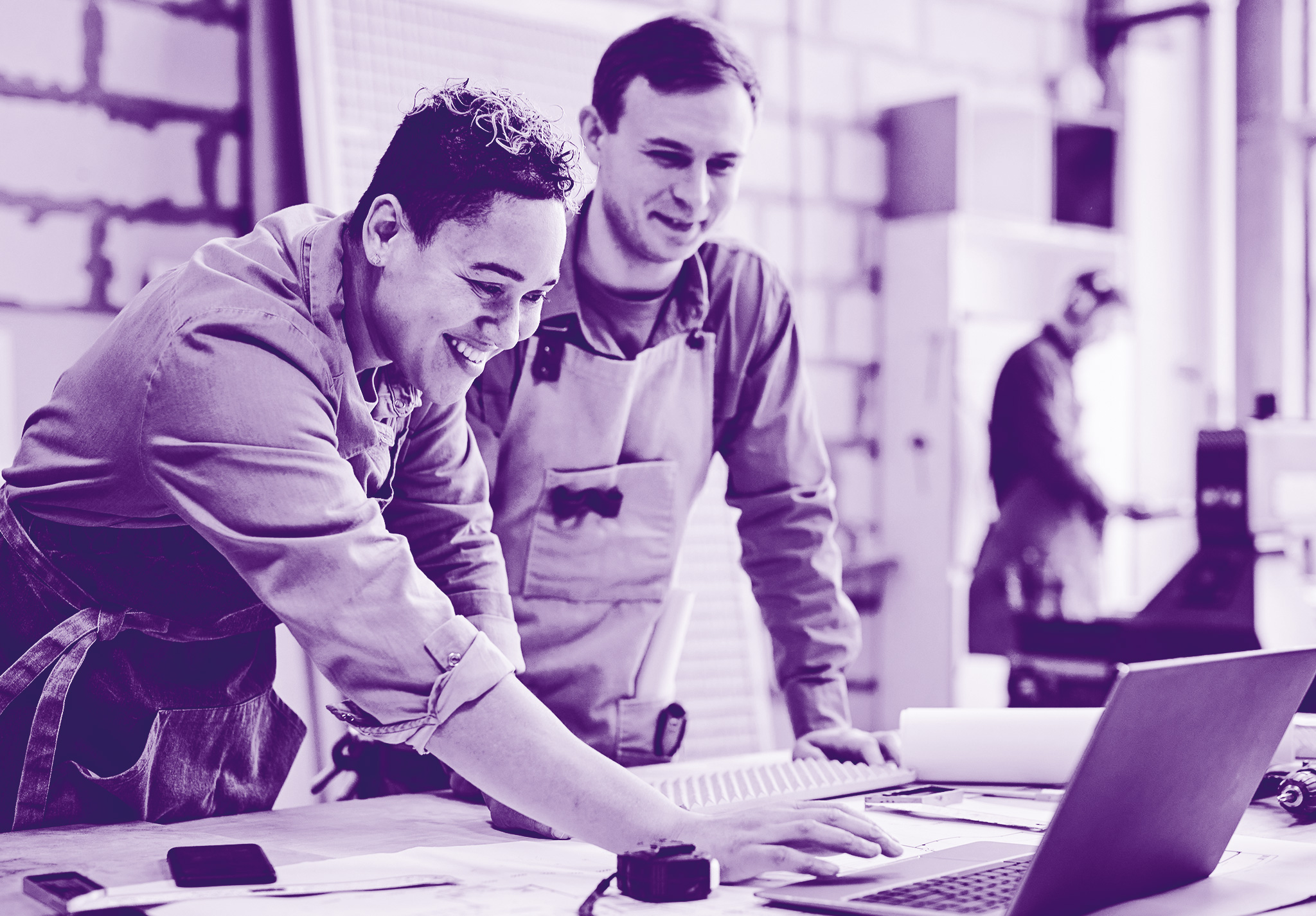 foto con sfondo sul viola con con due uomini vestiti da lavoro, appoggiati ad un tavolo che stanno guardando un pc e sorridono