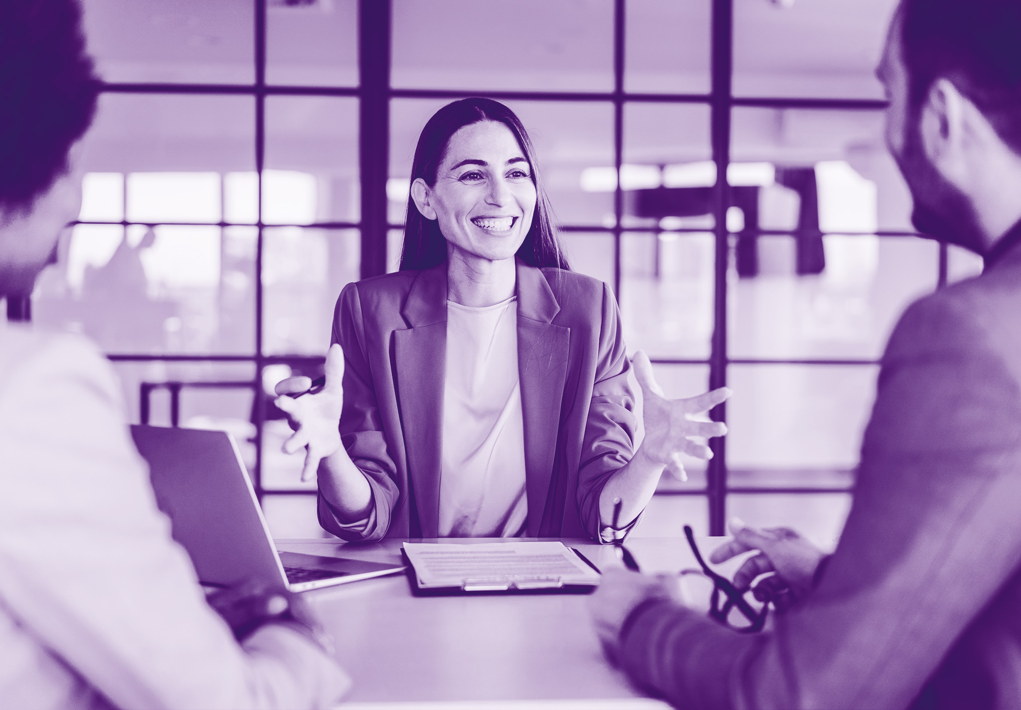 foto con sfondo viola con due persone in primo piano di spalle e una donna con i capelli lunghi in mezzo seduta alla scrivania che sorride, con davanti un pc e dei foglia, le braccia davanti a se con le mani aperte