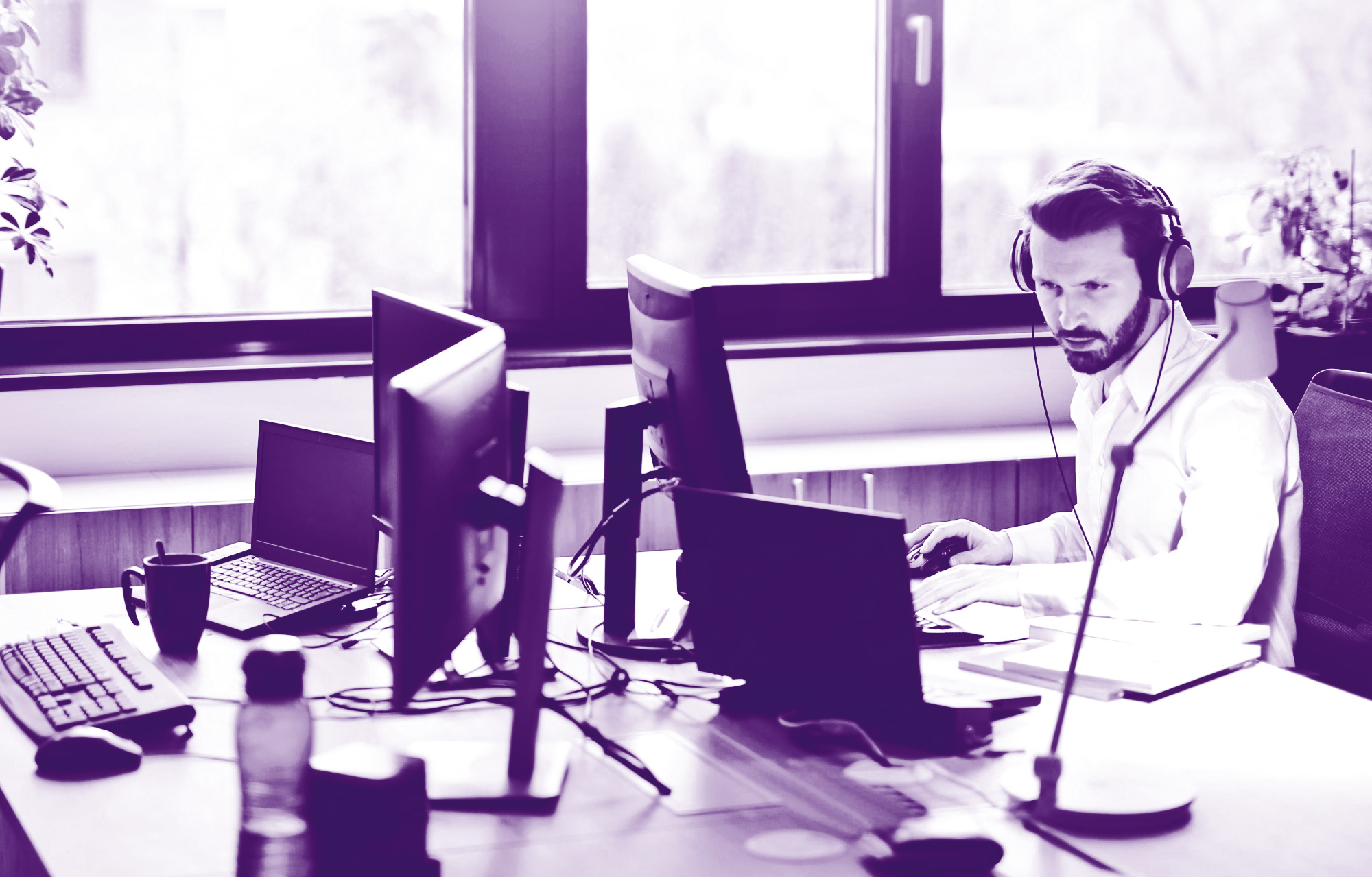 foto con gradiente viola che ritrae una scrivania con due pc e in uno c'è un ragazzo che lavora con le cuffie